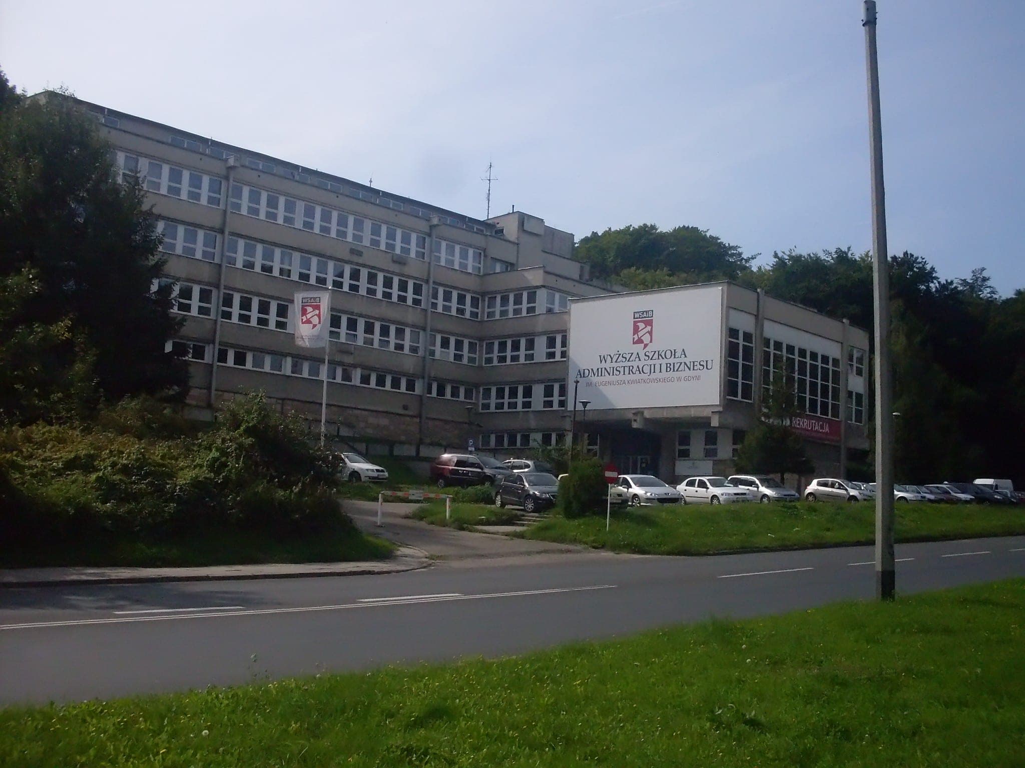 Yönetim ve İşletme Yüksekokulu adı yazılı, düzenli yerleştirilmiş pencereli çok katlı modernist bina, ön tarafta otopark alanı.